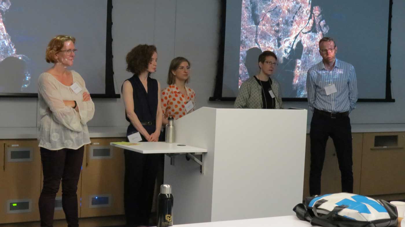 five speakers stand in front of a podium speaking to the audience