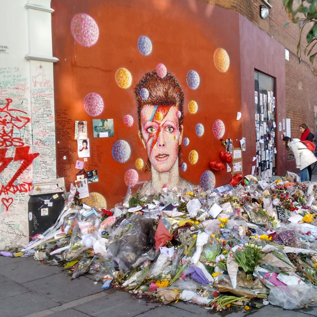 wall mural of David Bowie with pile of flower bouquets on the ground in front of it