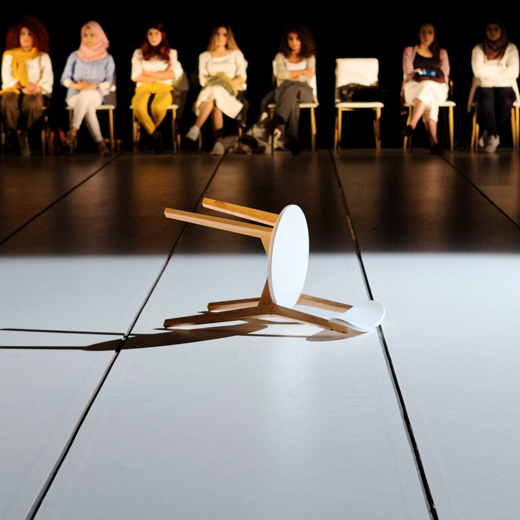 a row of people sit on chairs at rear of stage on which a chair lays on its side