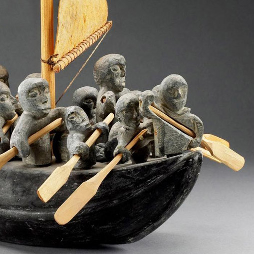 Photograph of sculpture made of gray stone and mixed materials portraying people on a boat with a gold sail and oars.