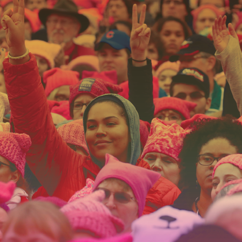 pink-toned photograph of a crowd of people, some with their arms up making peace signs and others wearing pink hats
