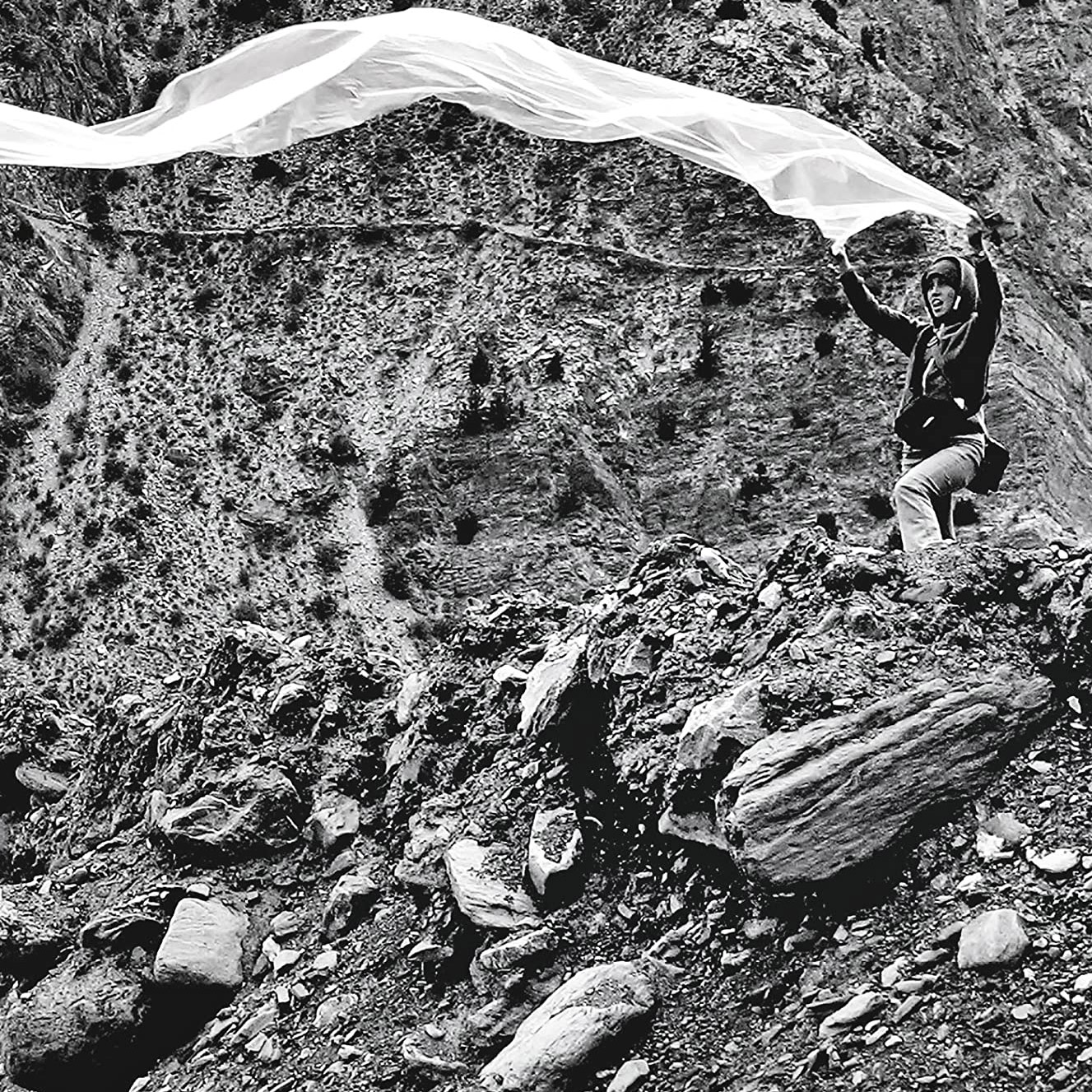 black and white image of a figure holding a long, white piece of fabric against a rocky background