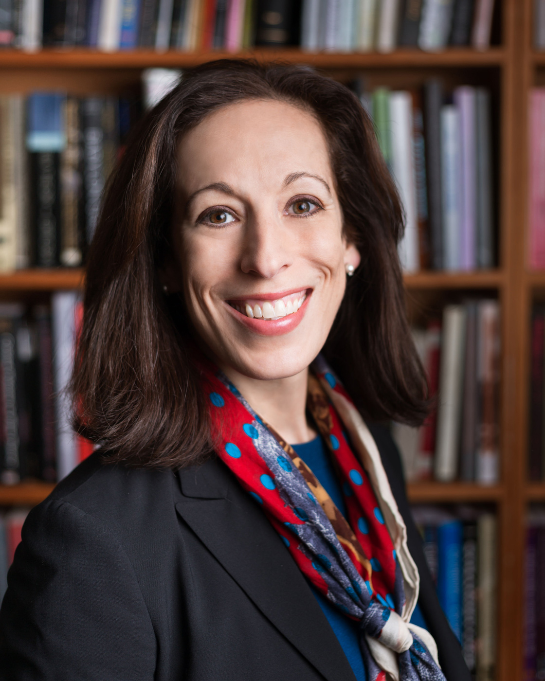 A person with shoulder-length hair and a brightly colored scarf stands smiling in front of a shelf of books