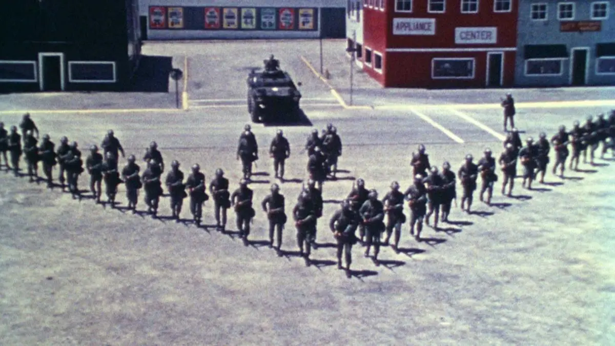 Still image from documentary film Riotsville, U.S.A. Police officers in riot gear in an arrow formation with a tank behind them.