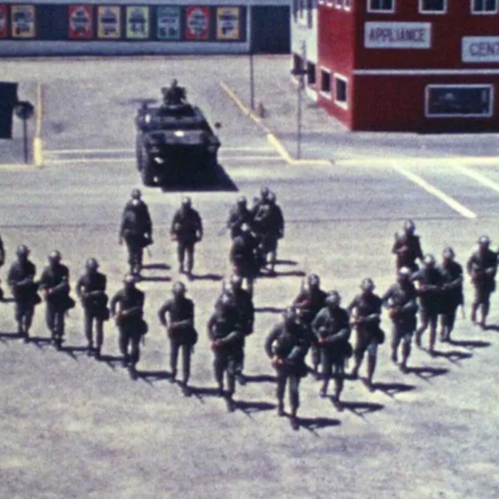Still image from documentary film Riotsville, U.S.A. Police officers in riot gear in an arrow formation with a tank behind them.